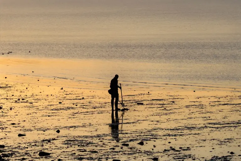 Silhouette einer Person mit Metalldetektor am Strand bei Sonnenaufgang